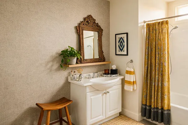 A warm eclectic master bathroom with a white vanity and integrated sink beneath an ornate carved wood mirror. A mustard patterned shower curtain, woven rug, wood stool, and small greenery add texture and color to the neutral room.