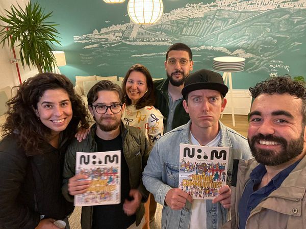 A group of six people poses together, holding magazines and smiling in a cozy indoor setting.