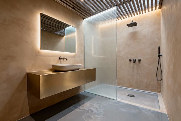 A modern bathroom with warm beige plaster walls features a floating brushed-brass vanity holding a pale stone vessel sink and a wall-mounted spout beneath a large, backlit mirror. A walk-in shower is defined by a reeded-glass screen, a ceiling-mounted square rain head, and matte black controls with a handshower; a slatted wood ceiling with concealed LEDs casts a soft glow over a seamless gray resin floor and integrated shower tray.