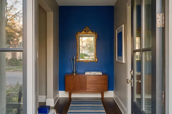 A narrow entryway with a vivid royal blue accent wall, a gold ornate mirror centered above a mid-century wooden console, and a striped blue rug on dark wood floors. Gray walls, white trim, and glass-paneled doors frame the space, with a small decorative sculpture and tray on the console.