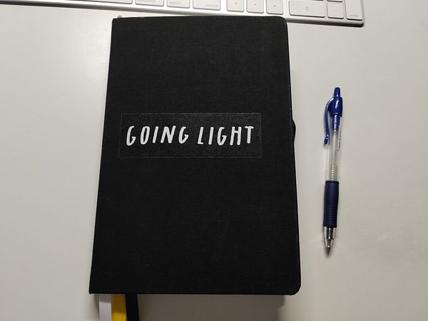 A black journal titled 'GOING LIGHT' is placed next to a blue pen on a white surface.