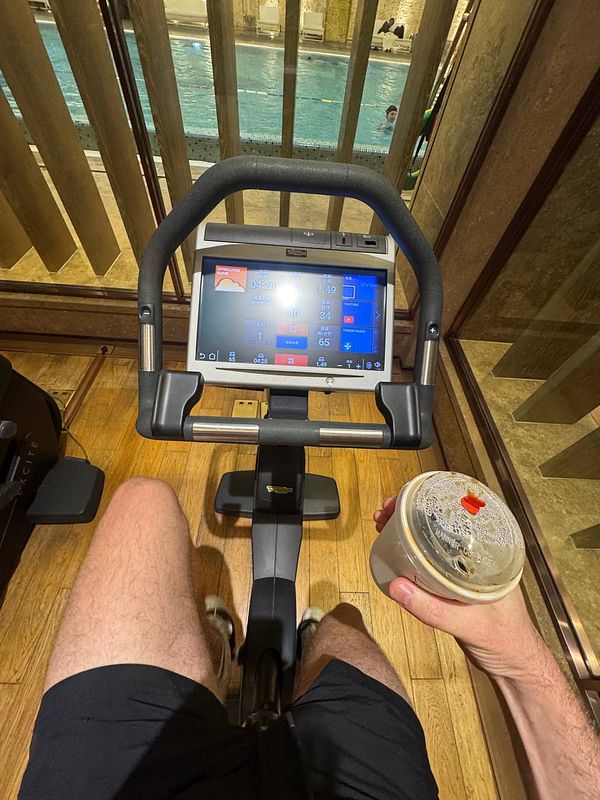 A person is using a stationary bike in a gym with a view of a swimming pool.