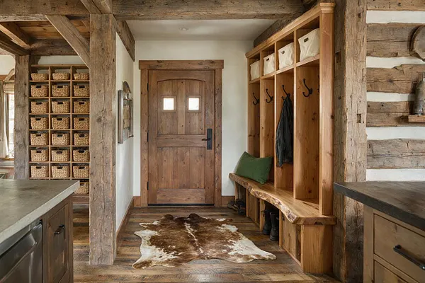 A rustic entryway with a paneled wooden front door, built-in wood cubbies and hooks, and a long bench with storage. A cowhide rug lies on the wood floor, while exposed timber beams, log walls, and woven baskets add texture and warmth.
