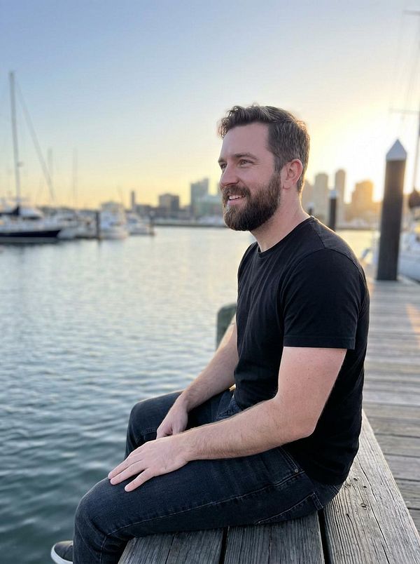 A man sits on a wooden dock by the water, enjoying a sunset view of the city skyline.