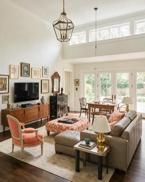 A bright eclectic game room with high ceilings, large windows, and French doors bringing in natural light. The room features a tan leather sectional, coral upholstered armchair, patterned ottoman, antique-style wood furniture, a small game table, and layered wall art with mixed vintage accents.
