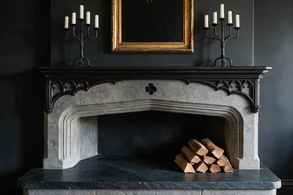 A dark Gothic-style fireplace vignette with a carved stone surround, black mantel, and a stack of firewood inside the hearth. Two black metal candelabras with white candles sit symmetrically on the mantel beneath a gold-framed empty picture frame against a deep charcoal wall.