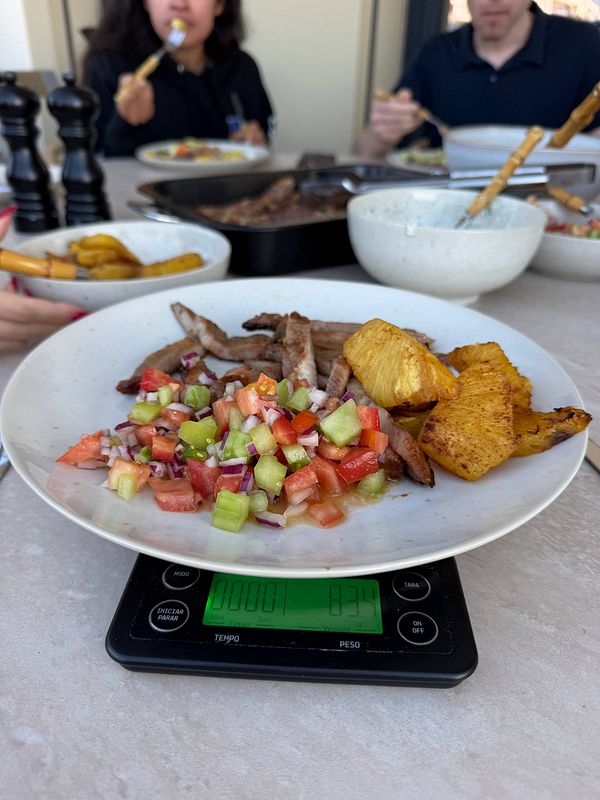Plate with grilled pineapple, salad, and meat on a digital kitchen scale during a group meal.