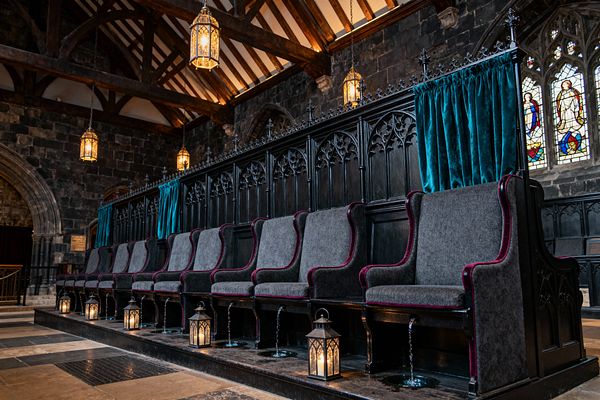 A dramatic media room styled like a chapel, with a long row of high-back gray seats trimmed in burgundy, built into black carved wood paneling with pointed-arch tracery. Teal velvet curtains, candlelit floor lanterns, and brass pendant lights illuminate dark stone walls, exposed timber beams, arched openings, and stained-glass windows over a checkerboard stone floor.