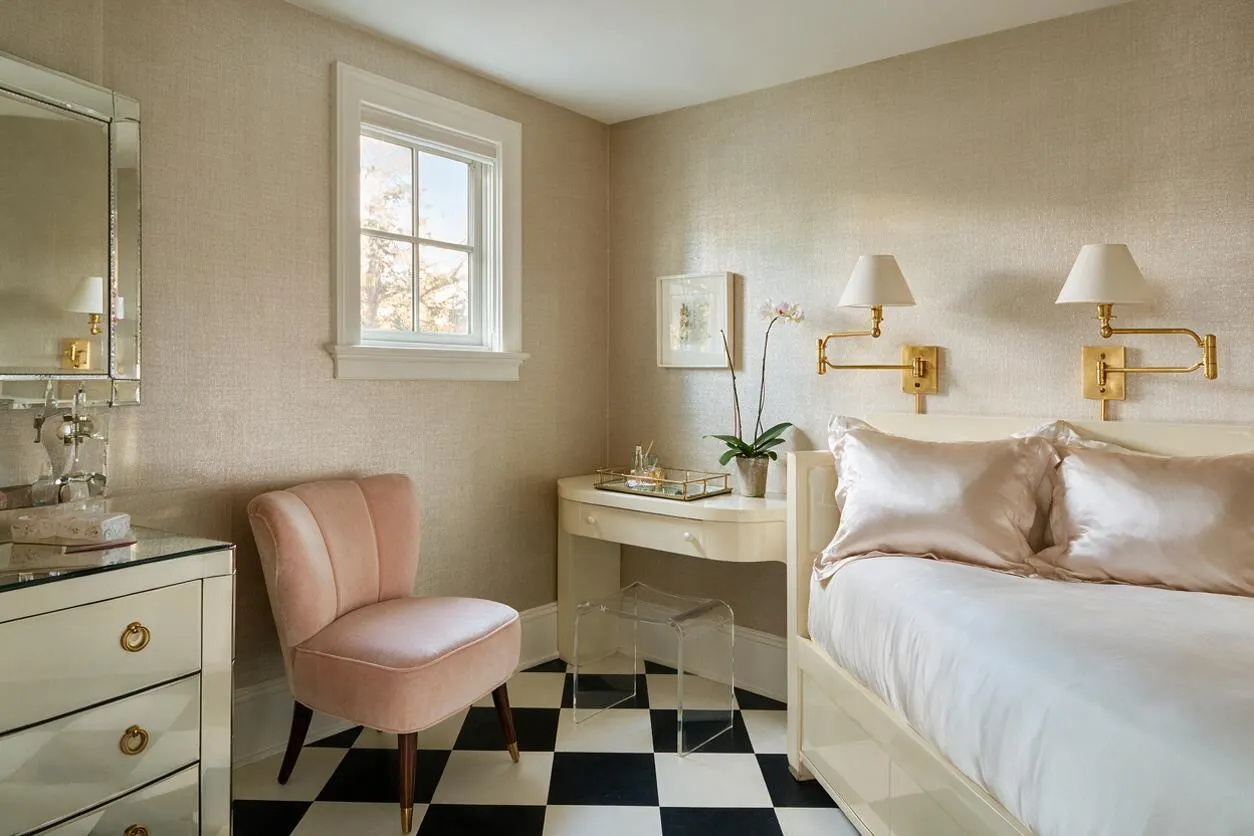 A stylish guest bedroom with a cream bed dressed in white bedding and blush satin pillows, paired with gold swing-arm wall lamps. The room features a black-and-white checkered floor, a pink velvet accent chair, a mirrored dresser, and a small cream desk under a window, creating a polished Hollywood Regency look.