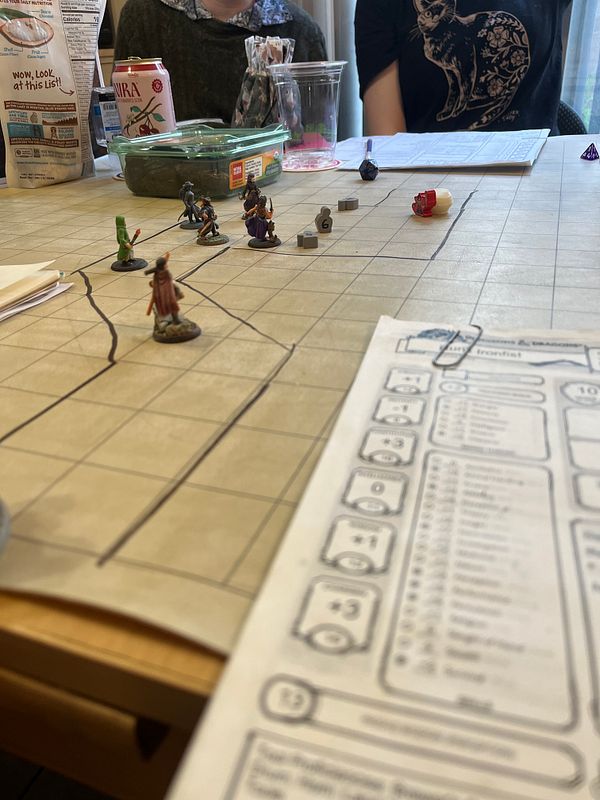 A tabletop scene from a Dungeons & Dragons game featuring miniatures, a character sheet, and various game materials.