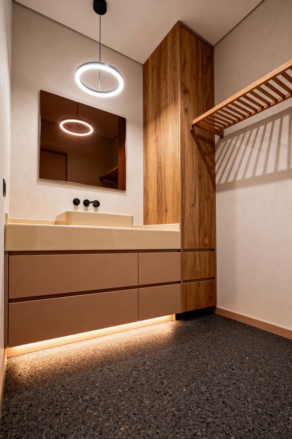 A compact modern laundry room with a floating clay-toned vanity and integrated cream utility sink, a wall-mounted matte black faucet, and a simple rectangular mirror. A circular LED ring pendant hangs above, with toe-kick lighting washing the floor. To the right, a tall vertical-grain wood cabinet and matching slatted drying shelf add storage, while warm sand-colored walls and a dark speckled terrazzo floor complete the minimal, clean look.