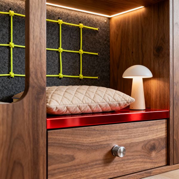 Close-up of a modern children’s built-in cubby made of walnut. A charcoal felt back panel holds a grid of neon-yellow rope secured with small hardware. Warm LED strip lighting lines the top, illuminating a glossy red metal shelf with a small mushroom-shaped table lamp and a quilted beige cushion. Beneath the shelf is a walnut drawer with a knurled stainless knob. Curved cutouts soften the edges, creating a cozy, playful reading nook.