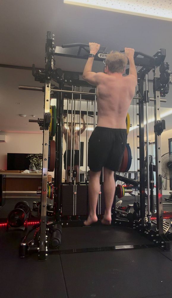 A young man performing pull-ups on a yellow and blue outdoor gym structure in a park.