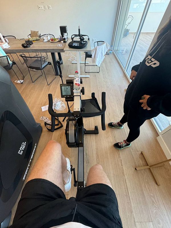 A person is using a rowing machine in a home gym setting with a personal trainer nearby.