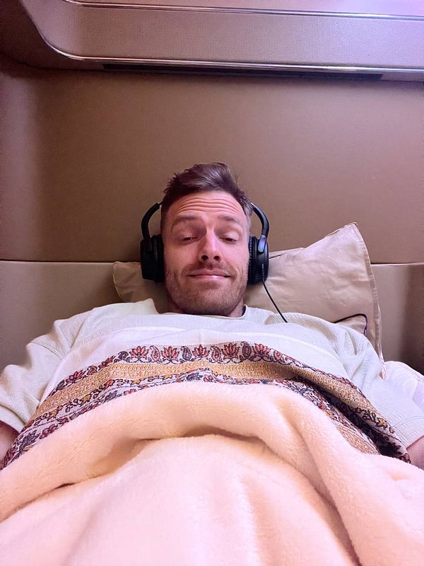 A relaxed individual is lying down in a luxurious airplane cabin, wearing headphones and covered with a blanket.