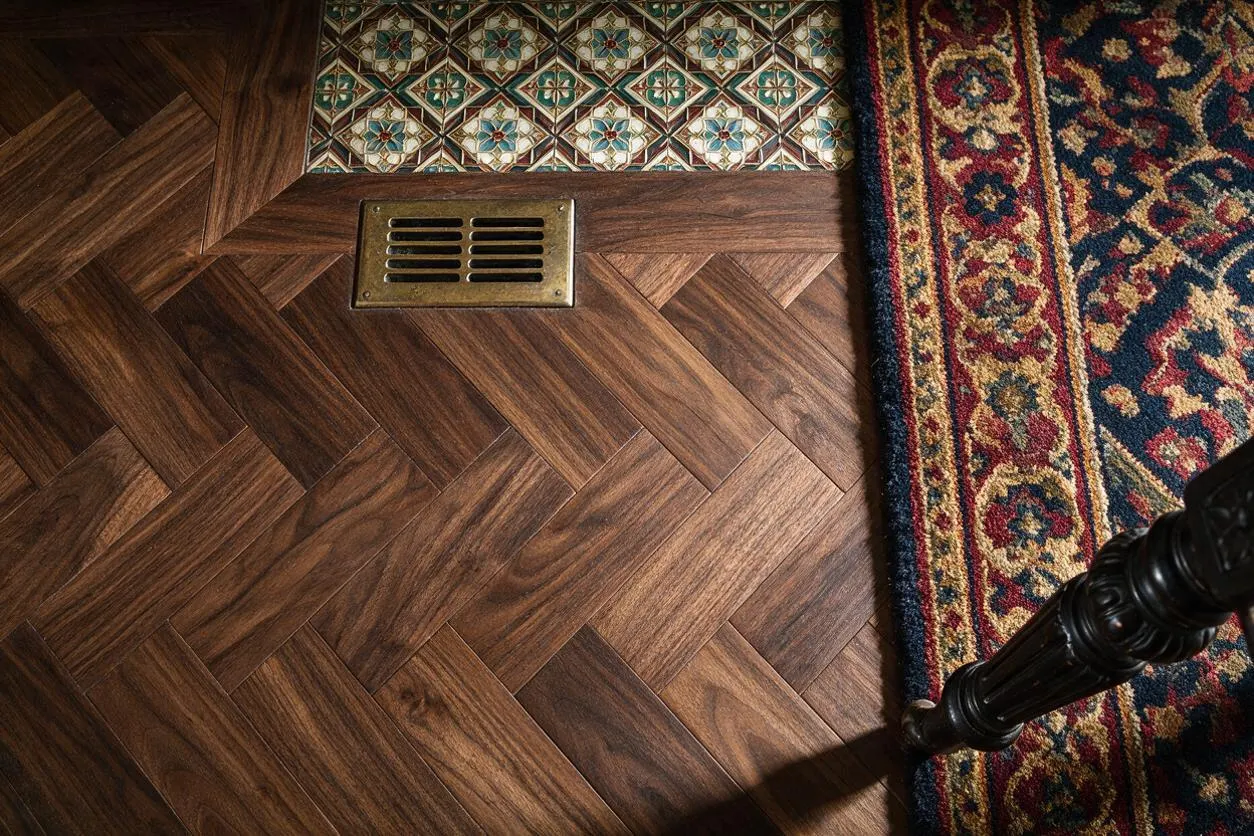 A close-up view of a Victorian-style interior floor featuring rich herringbone wood planks, a brass floor vent, a strip of decorative patterned tile, and the edge of a traditional Persian-style rug. Dark carved furniture legs appear at the lower right, adding to the classic period feel.