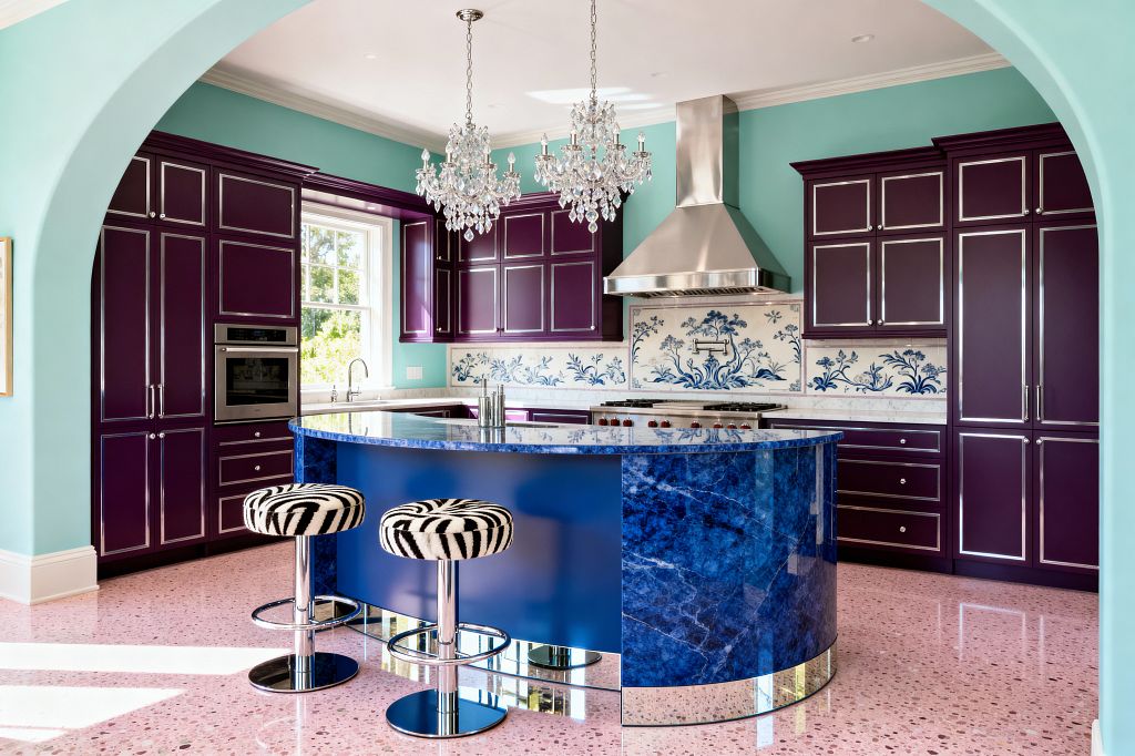 A Hollywood Regency kitchen with glossy plum cabinets edged in chrome against aqua walls. A stainless-steel range hood crowns a professional range backed by a white tile backsplash with blue floral motifs. At center, a curved island clad in cobalt blue stone features a mirrored toe-kick and a dark polished top. Two zebra-print stools on chrome bases sit on a pink terrazzo floor, while twin crystal chandeliers hang above. An arched opening frames the room and daylight pours through the window.