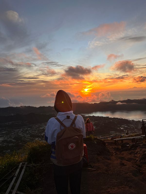 A person stands on a mountain overlooking a stunning sunrise with clouds and a view of the landscape below.