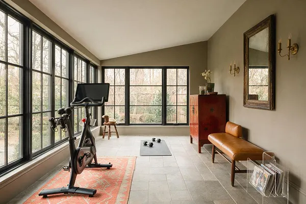 A bright home gym with large black-framed windows on two sides, a stationary bike on a patterned rug, and a yoga mat with dumbbells in the center. The room mixes workout equipment with eclectic decor, including a leather bench, a red vintage cabinet, a large wall mirror, brass sconces, and a clear magazine rack.