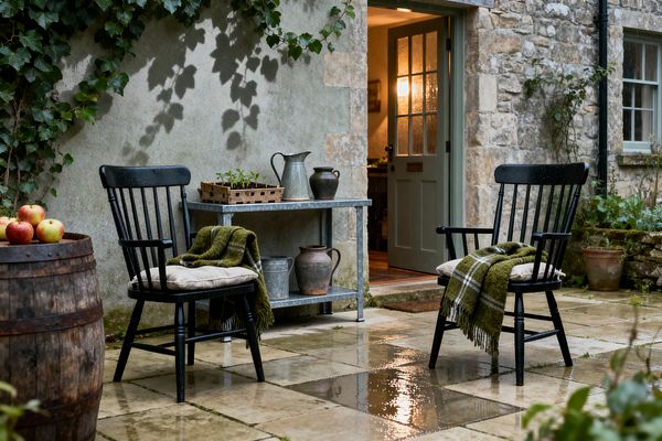 A rain-wet stone patio outside a rustic cottage features two black spindle-back chairs with beige cushions and green plaid throws. A galvanized metal potting table holds a zinc watering can, clay jugs, and a crate of seedlings. Ivy climbs a textured plaster wall beside a rough stone façade. An open sage green door glows with warm interior light, while a weathered wooden barrel topped with apples and terracotta planters complete the scene.