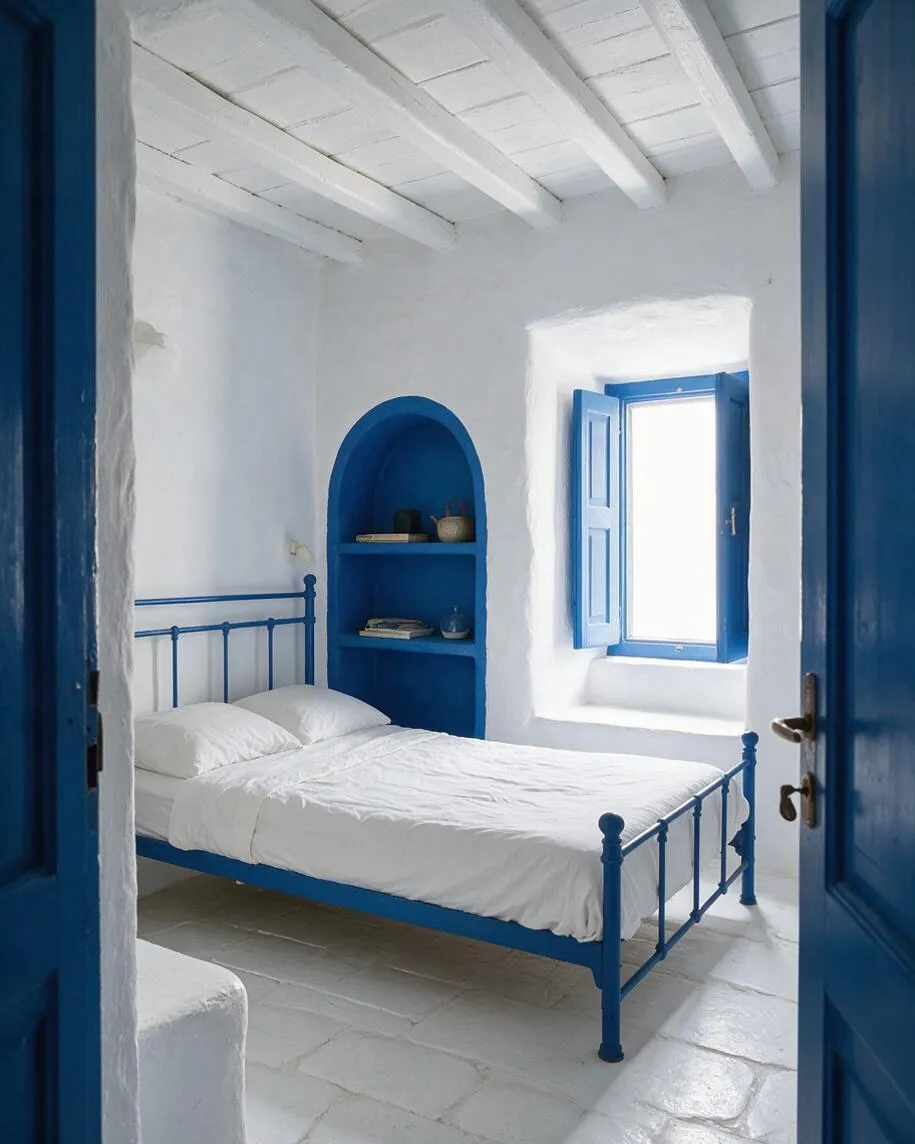 A compact basement bedroom with white plaster walls, exposed white ceiling beams, and a vivid blue metal bed frame. Built-in arched shelving and a blue-shuttered window add Mediterranean character, while the stone-like floor and bright natural light keep the room airy.