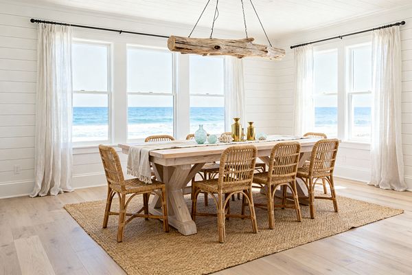 An airy coastal dining room with white shiplap walls and four large windows overlooking the ocean. A light wood trestle table seats eight rattan chairs on a woven jute rug, topped with a neutral runner, brass candlesticks, sea-glass vases, and shells; sheer white linen curtains on black rods frame the view as a suspended driftwood beam pendant centers the space, with pale wood plank flooring and abundant natural light.