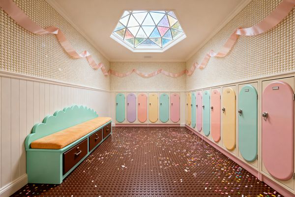 A whimsical mudroom inspired by Candy Land with two rows of cream lockers fitted with rounded pastel doors in mint, blush, peach, lemon, and lavender. A scalloped mint bench with a caramel cushion and brown drawers lines one wall; above, beadboard wainscoting transitions to pearly bubble tile draped with pink ribbon, while a geometric stained-glass skylight brightens a chocolate-brown coin rubber floor scattered with confetti-like sprinkles.