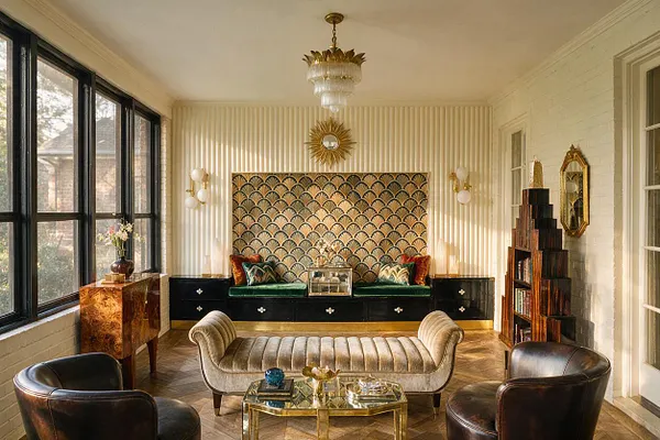 A sunlit front porch styled in Art Deco fashion with black-framed windows, cream walls, and a gold fan-patterned feature wall. The space includes a green velvet built-in bench, a ribbed cream settee, two dark leather chairs, glass-and-gold tables, brass wall sconces, a sunburst mirror, and a tiered chandelier.