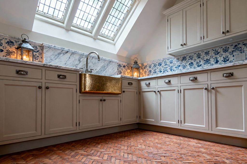 A traditional laundry room features greige inset cabinets with antique nickel hardware, Carrara marble countertops and backsplash, and a hammered brass apron-front sink. A band of blue-and-white Delft-style tiles lines the wall, while a herringbone terracotta brick floor adds warmth. Three leaded-glass skylights bring in natural light, and two candlelit metal lanterns sit on the counter for a soft, ambient glow.