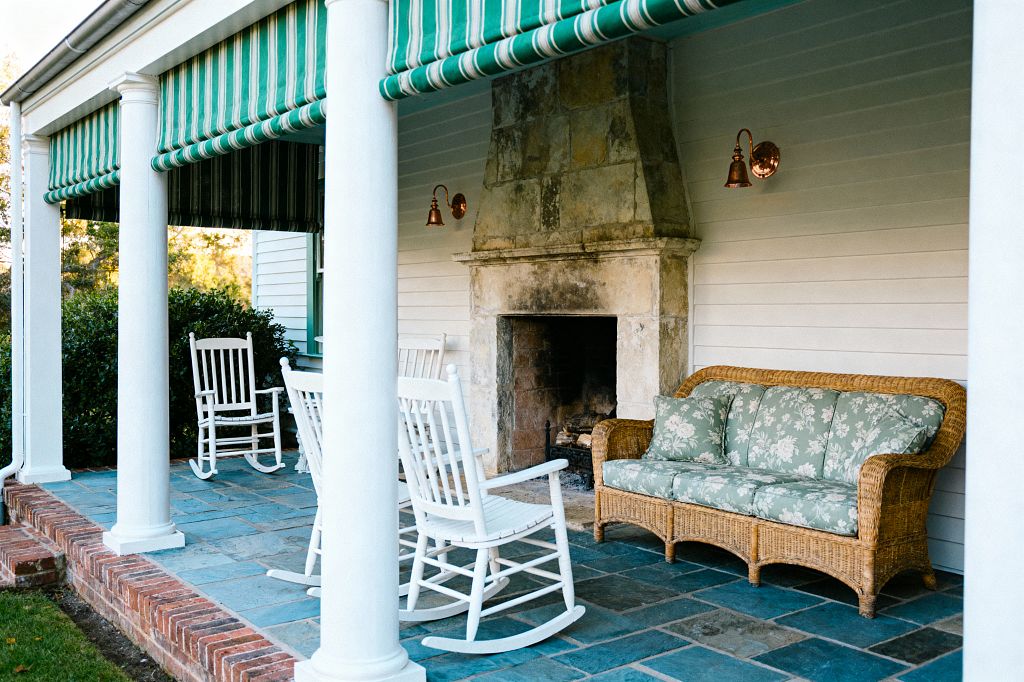 A covered traditional porch features round white columns supporting a roof shaded by green-and-white striped awnings. A large, weathered stone fireplace anchors the space, flanked by copper wall sconces. Two white wooden rocking chairs sit on slate flagstone flooring near a wicker sofa with sage green floral cushions. White clapboard siding, a brick step, and neatly trimmed shrubs frame the patio, creating a calm, classic setting for relaxed outdoor living.