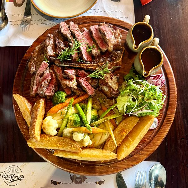 A wooden platter with sliced grilled steak, vegetables, fries, a fresh salad, and two small pitchers of sauce, set on a table for lunch.