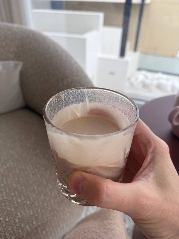 A hand holds a glass of beef protein shake in a cozy indoor setting.