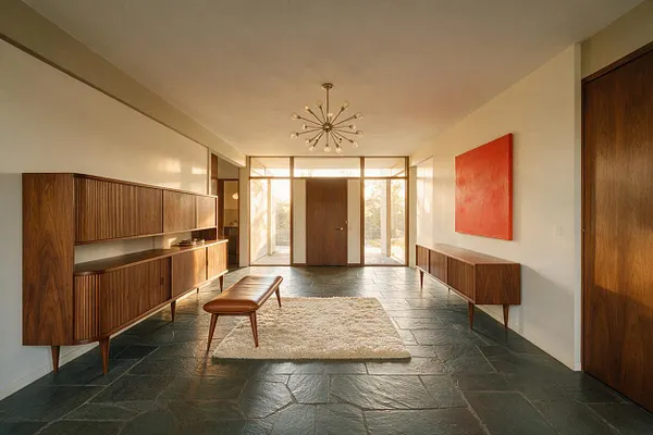 A spacious mid-century modern entryway with dark slate flooring, walnut storage cabinets on both sides, and a small leather bench centered on a cream shag rug. A starburst ceiling light hangs overhead, while a bright orange abstract painting adds color to the white wall near the front door.