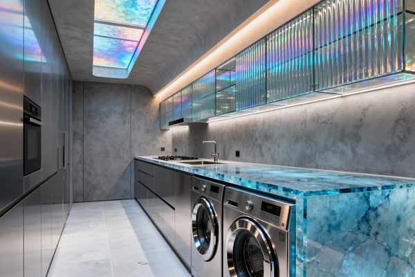 A sleek, futuristic laundry room features two front-loading stainless steel machines beneath a thick, backlit blue onyx countertop with a waterfall edge. Iridescent fluted-glass wall cabinets glow under linear LED strips above gray microcement walls and a curved ceiling. A prismatic ceiling panel casts rainbow hues across the space, complemented by tall stainless cabinetry and large-format light gray floor tiles.