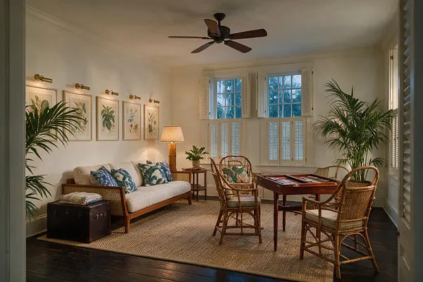 A tropical-style game room with a cream sofa, wicker chairs around a wooden game table, and large potted palms in the corners. Framed botanical prints, plantation shutters, a woven rug, and warm lamp light create a relaxed, airy atmosphere.