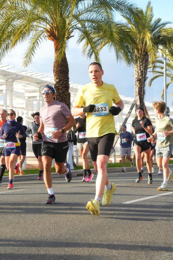 A runner in a green shirt and black shorts running downhill on a paved path in a forest during autumn.