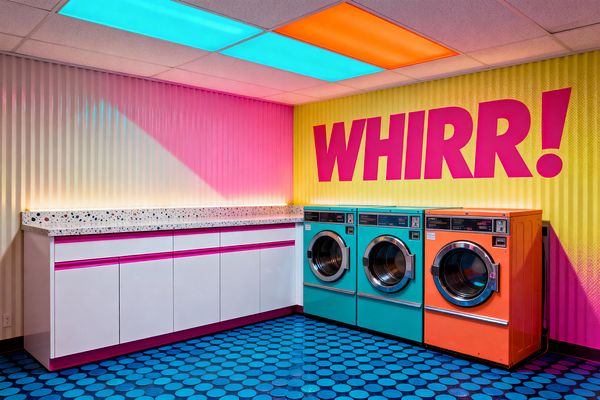 A Pop Art laundry room features three front-loading machines, two teal and one orange, set against a corrugated wall that fades from pink to yellow with a large magenta “WHIRR!” graphic. White cabinets with fuchsia trim support a chunky terrazzo countertop. Cyan and orange ceiling light panels and soft cove lighting create a neon glow, while a deep blue floor with lighter circular dots completes the playful scene.