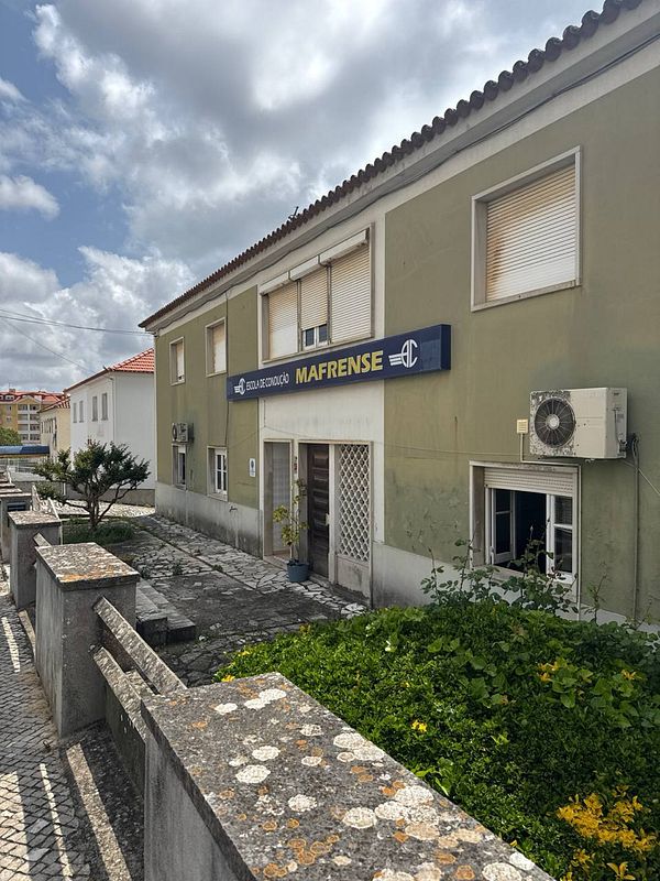 The image depicts a driving school building with a sign indicating its name and location.