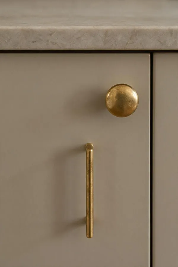 A close-up of a taupe cabinet door beneath a light stone countertop, featuring a rounded brass knob and a slim vertical brass pull. The composition highlights the smooth painted finish, warm metallic hardware, and clean Transitional styling.