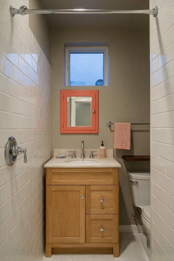 A narrow master bathroom with white subway tile walls, a light oak vanity topped with a pale stone countertop, and chrome fixtures. A coral-framed mirror adds a bold eclectic accent beneath a small high window, with a patterned towel and toilet visible to the right.