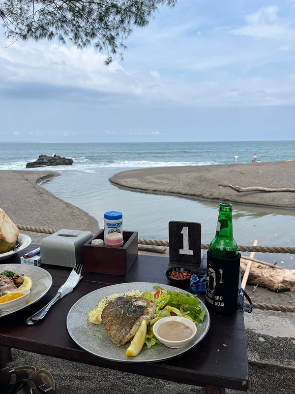 A scenic beachside lunch setup featuring a plate of grilled fish and a bottle of beer.