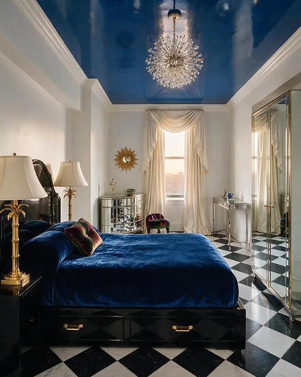 A glamorous guest bedroom features a deep blue velvet bed on a black base, mirrored furniture, and a sparkling starburst chandelier beneath a high glossy blue ceiling. Cream drapery frames the window, while black-and-white diamond tile flooring and gold accents create a dramatic Hollywood Regency look.