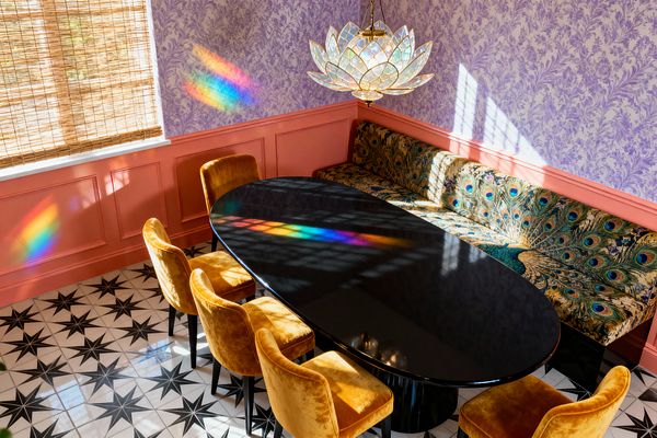 A maximalist dining room with a glossy black oval table surrounded by mustard velvet chairs and a long corner banquette upholstered in peacock-feather fabric. Lilac botanical wallpaper sits above coral-pink wainscoting, with black-and-white star tiles underfoot; woven bamboo shades filter sunlight that refracts from an iridescent lotus-shaped stained-glass chandelier, casting rainbow streaks across the wall and tabletop.
