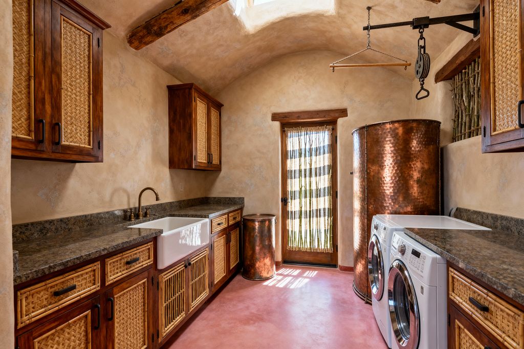 A Southwestern-style laundry room with hand-troweled beige plaster walls, a vaulted ceiling with exposed wood beams, and a skylight. Alder cabinets with woven cane panels line both sides, topped by dark granite countertops and a white farmhouse sink with a brass faucet. A rosy stained-concrete floor and hammered copper pieces, including a cylindrical tank and bin, add warmth. A white front-loading washer and dryer sit beneath the counters beside a wood-framed door with a reed screen.