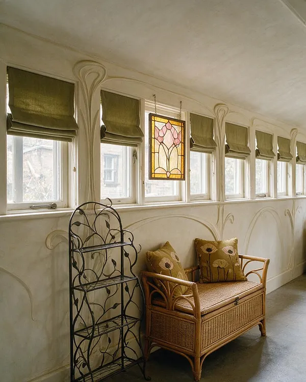 A narrow sunroom features a row of small windows with olive green Roman shades, sculpted plaster wall details, and a hanging stained glass panel in soft pink and amber tones. A wicker storage bench with patterned cushions sits beside a black metal vine shelving unit, creating a delicate Art Nouveau-inspired space.