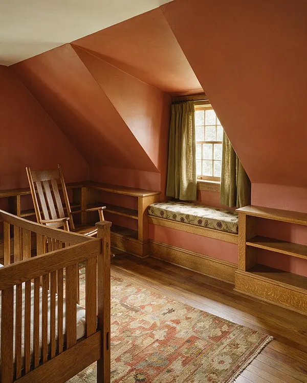 A Craftsman-style nursery with sloped terracotta walls, a wooden crib in the foreground, a wooden rocking chair, built-in shelving, and a cushioned window seat beneath a small window with green curtains. A patterned area rug and warm wood floors add softness to the cozy, attic-like room.