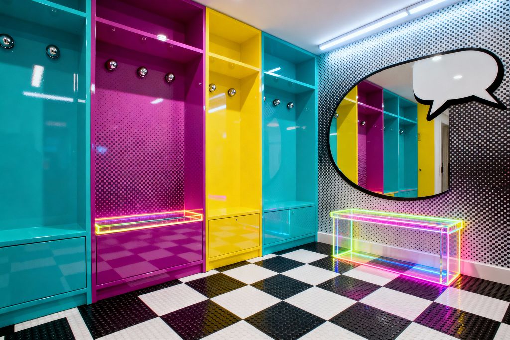 Color-blocked mudroom with glossy cyan, magenta, and yellow lockers, each with chrome hooks and bench drawers, set against walls printed with black-and-white halftone dots. A large oval mirror outlined as a comic speech bubble hangs above a clear acrylic bench that glows in neon rainbow edges. The floor is a black-and-white checkerboard with raised coin texture, while linear LED lights brighten the saturated surfaces and reflective finishes.