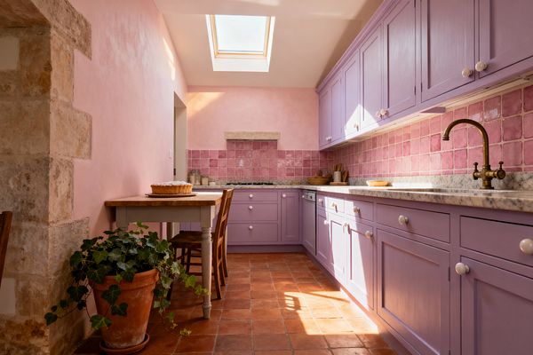 A sunlit galley kitchen features lavender Shaker cabinets with white marble countertops and a pink square-tile backsplash. Blush limewashed walls, terracotta floor tiles, and an antique brass bridge faucet add vintage warmth. A small farmhouse table with wooden chairs sits to the left, topped with a cake, beside a terracotta pot of ivy. A stone doorway and a stone shelf above the cooktop and a ceiling skylight complete the airy, Shabby Chic setting.