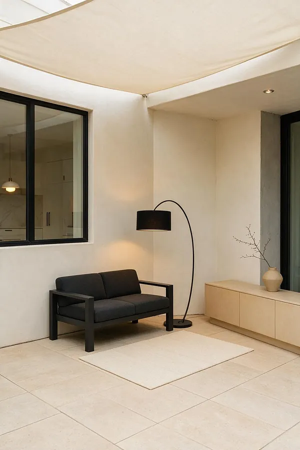 A minimalist backyard patio with large beige tile flooring, smooth white walls, and a stretched light canopy overhead. Black-framed windows and a slim black arched floor lamp anchor a small seating area with a dark sofa, a cream rug, and a light wood cabinet with a vase and branches.