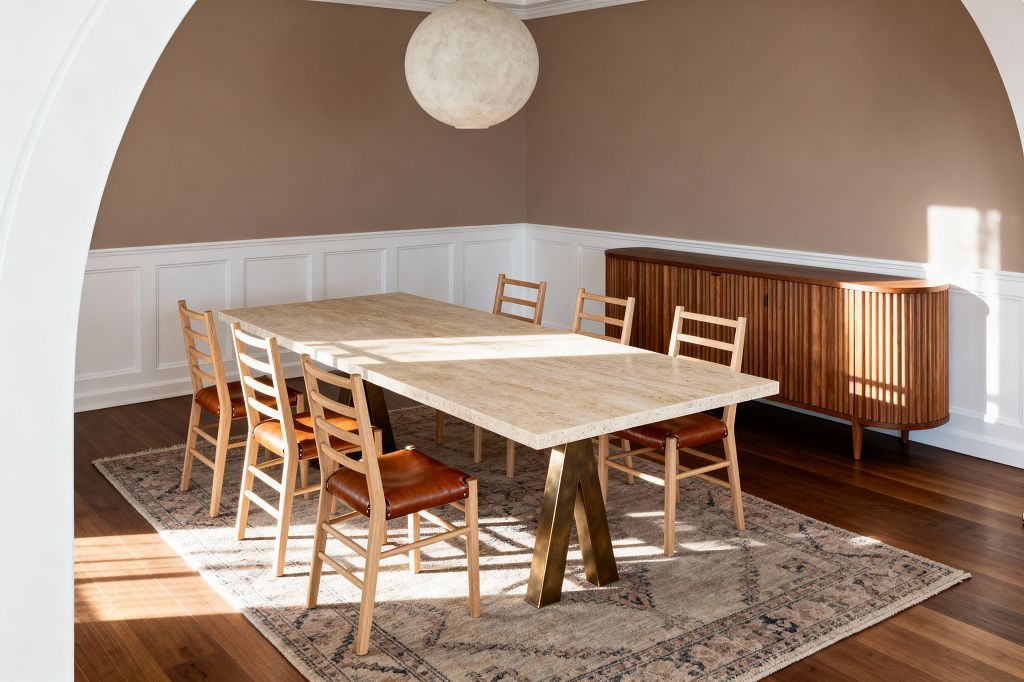 A transitional dining room framed by a white arch features a rectangular travertine table on sculptural brass legs, surrounded by six light oak ladder-back chairs with cognac leather seats atop a muted vintage-style rug. Warm taupe upper walls, white paneled wainscoting and crown molding, a fluted walnut credenza, medium-tone wood floors, and a textured globe pendant complete the scene with sunlight casting long shadows.